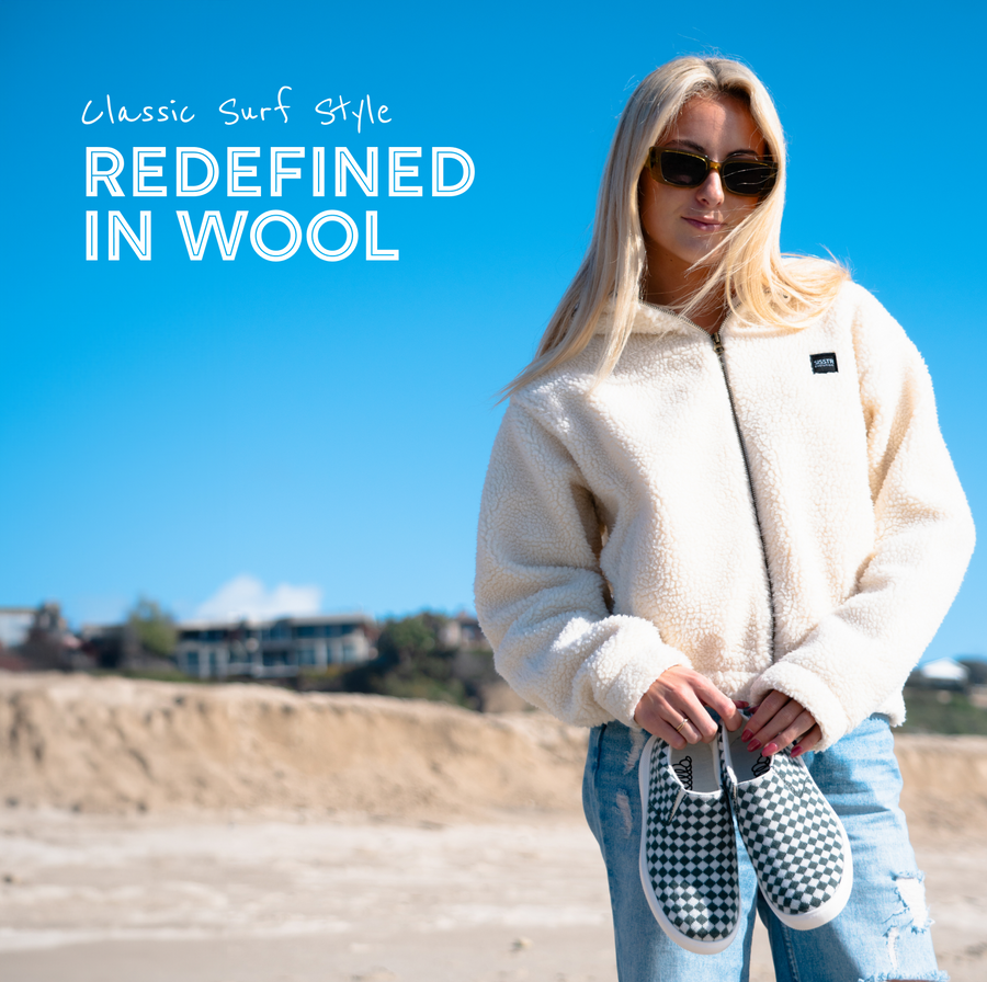 Woman wearing sunglasses and a cream fleece jacket holding black-and-white checkered Woolloomooloo slip-on sneakers, standing on a beach with text reading 'Classic Surf Style Redefined in Wool