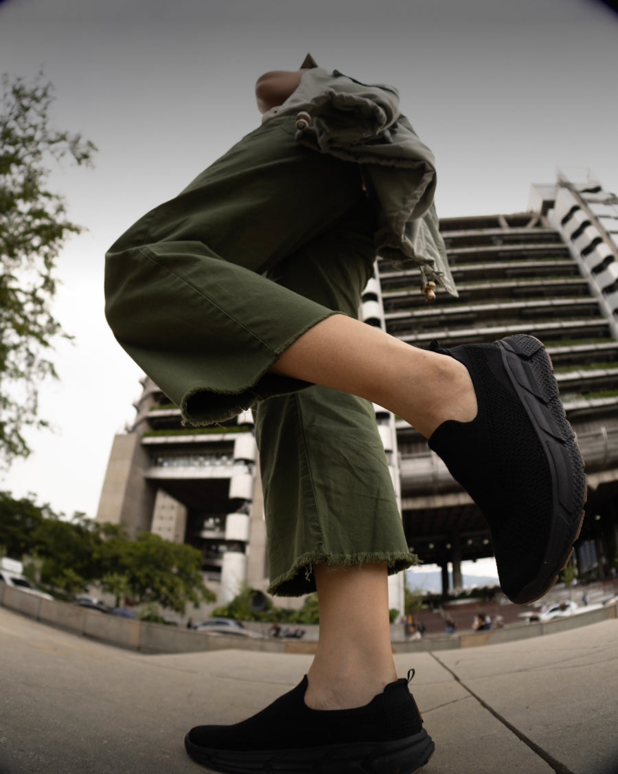 Low-angle view of a person walking in an urban setting wearing black Woolloomooloo slip-on sneakers and green cropped pants
