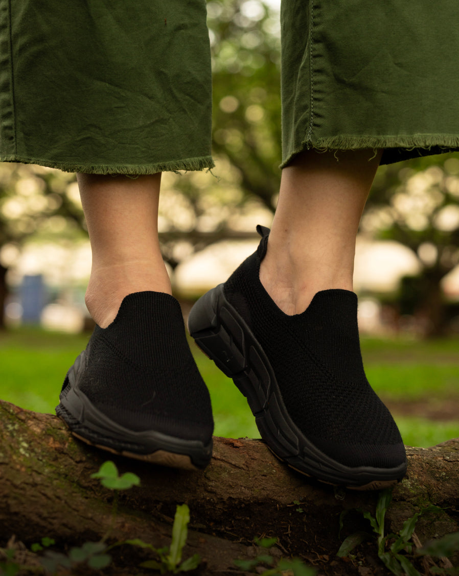 Close-up of feet wearing black Woolloomooloo slip-on sneakers with chunky black soles, standing on a tree branch in an outdoor park setting