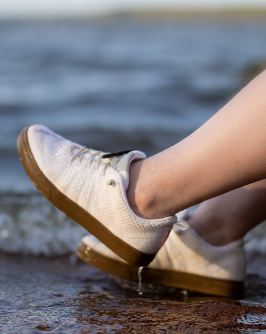 Close-up of feet wearing white Woolloomooloo lace-up sneakers with brown soles, resting at the edge of a shoreline with water splashing