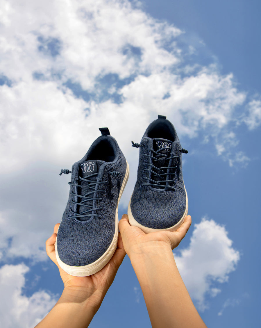 Hands holding a pair of navy blue Woolloomooloo lace-up sneakers with white soles against a bright blue sky with clouds.