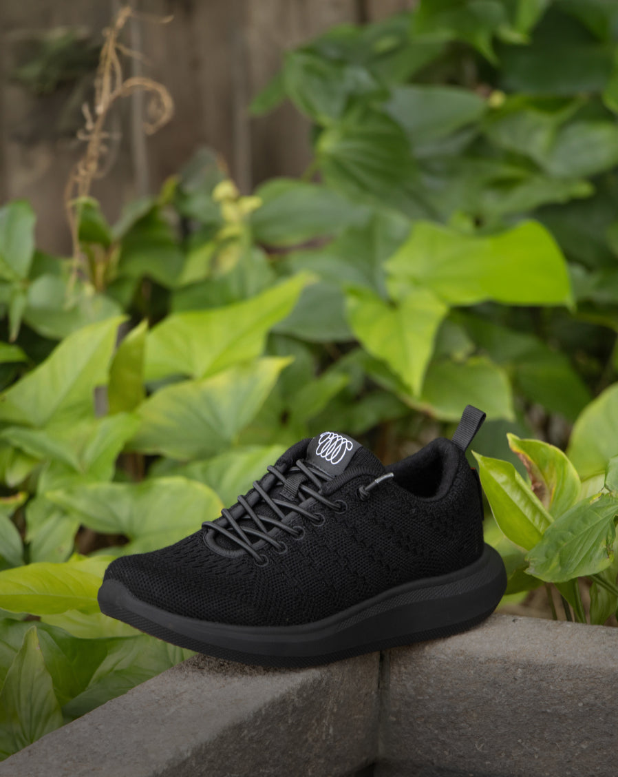 Black Woolloomooloo lace-up sneaker with matching black sole displayed outdoors on a stone surface with green leafy plants in the background