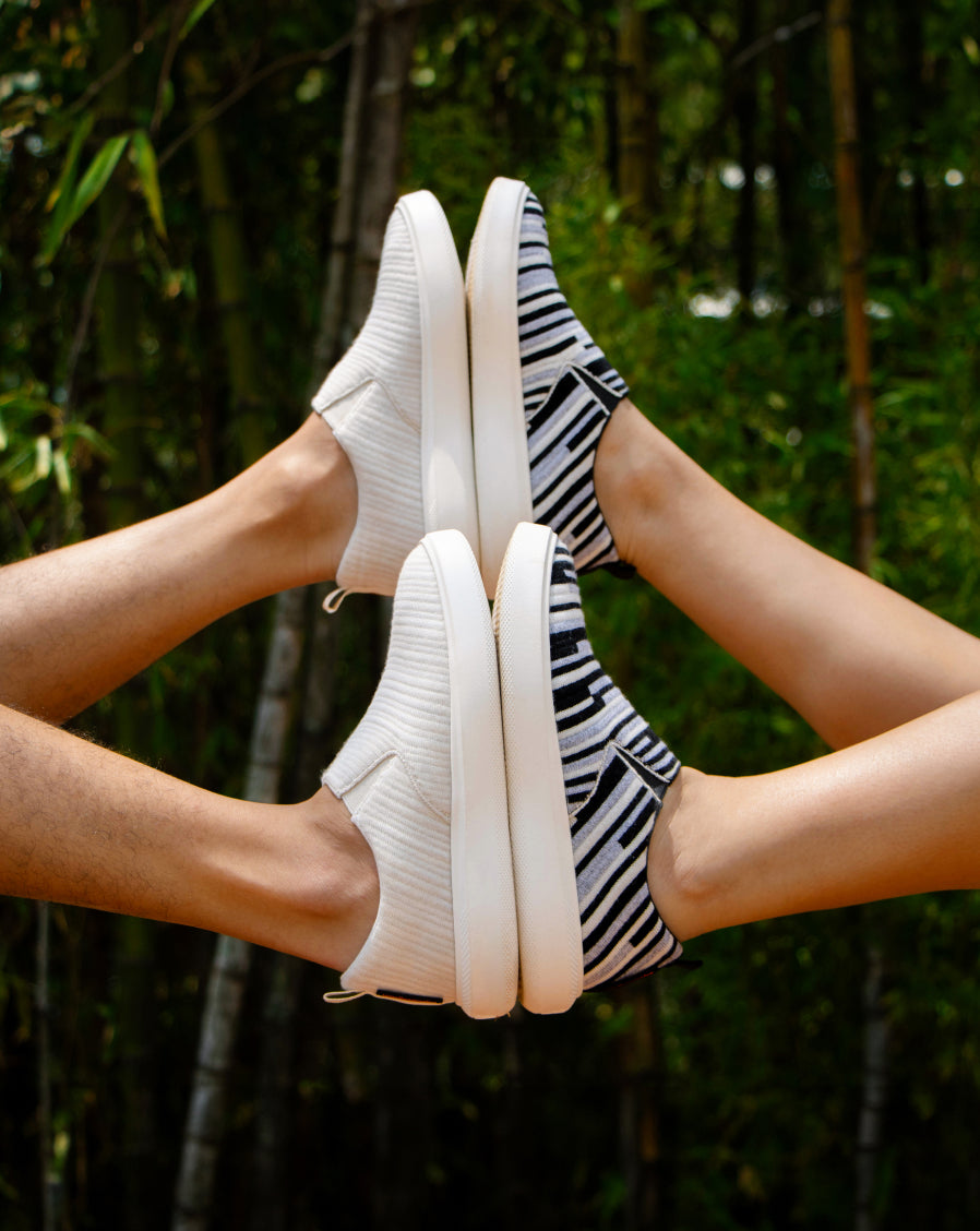 Two pairs of legs raised in the air, each wearing Woolloomooloo slip-on sneakers—one in solid white and the other in black-and-white stripes.