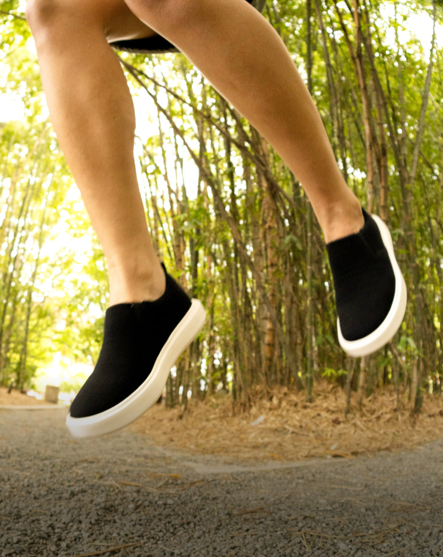 Person jumping on a forest trail wearing black Woolloomooloo shoes,