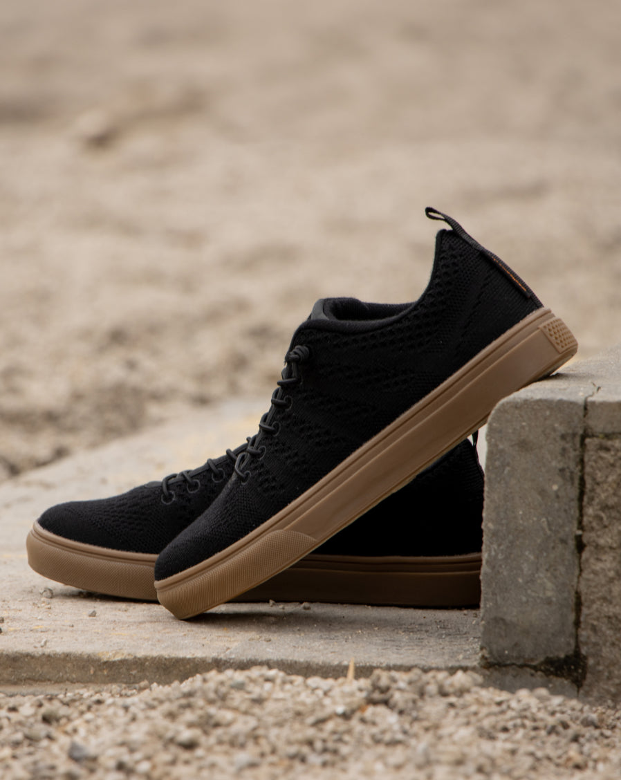 Black Woolloomooloo lace-up sneakers with brown soles, displayed outdoors resting against a concrete block