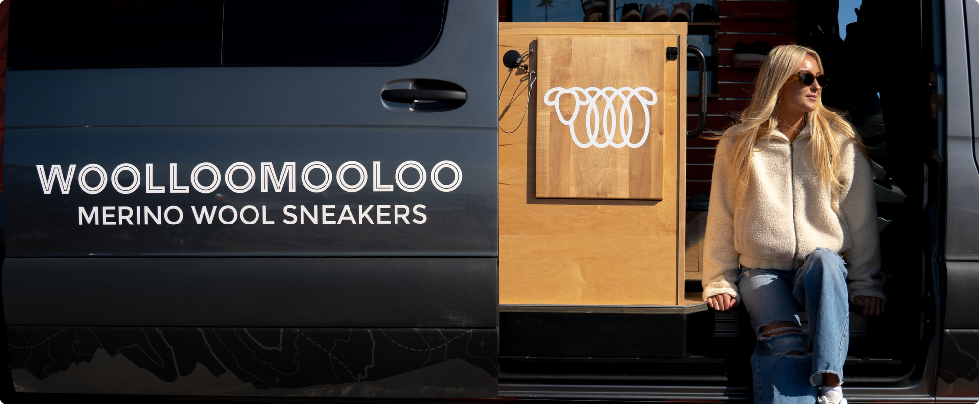 Woman in sunglasses and a cream fleece jacket sitting at the open door of a black Woolloomooloo van with 'Woolloomooloo Merino Wool Sneakers