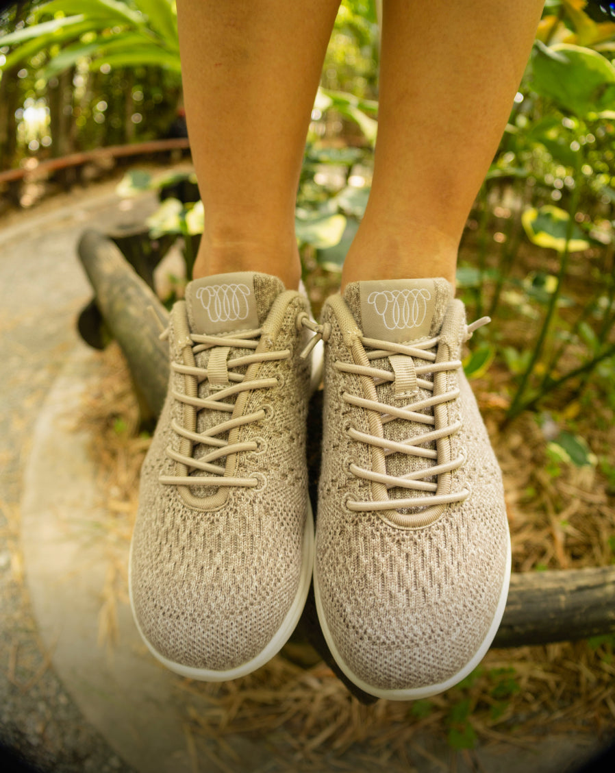 Close-up of feet wearing beige Woolloomooloo lace-up sneakers with white soles, standing on a wooden rail in a lush green outdoor setting.