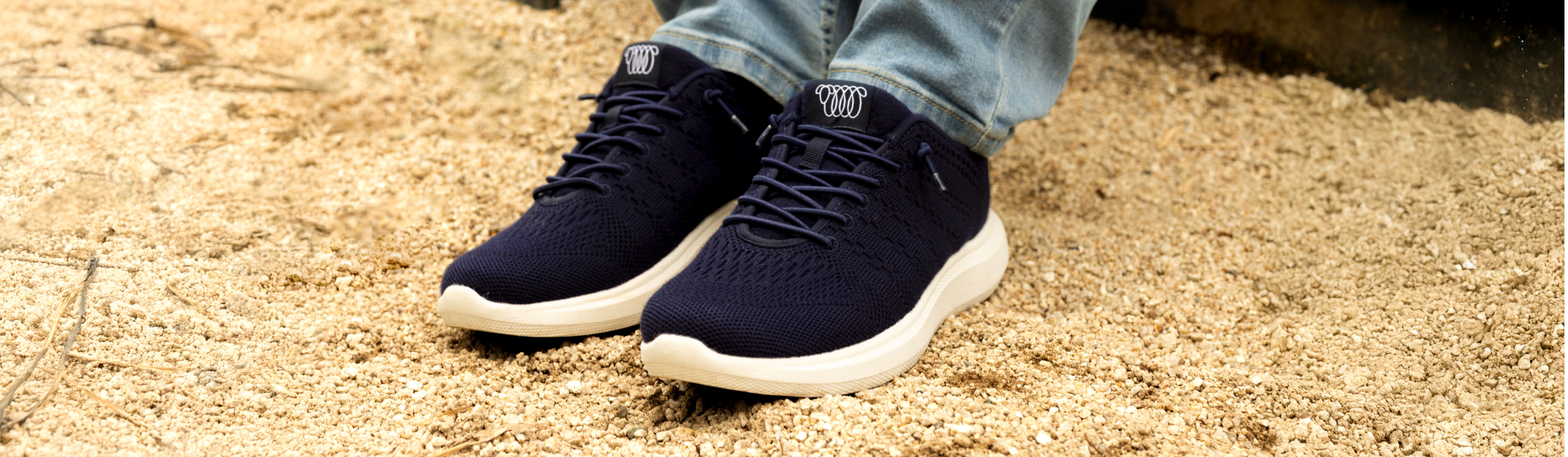 Close-up of person wearing navy blue Woolloomooloo sneakers with white soles, standing on a gravel surface in light blue jeans