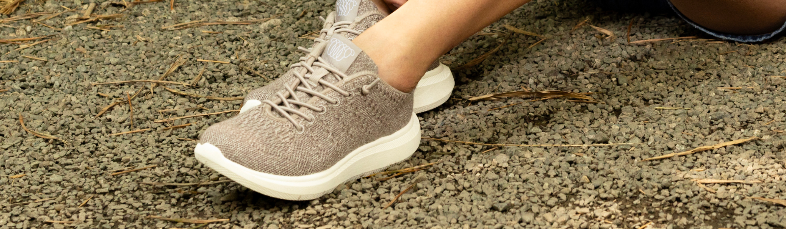 Close-up of person wearing Beige Woolloomooloo sneakers with white soles, standing on a gravel surface